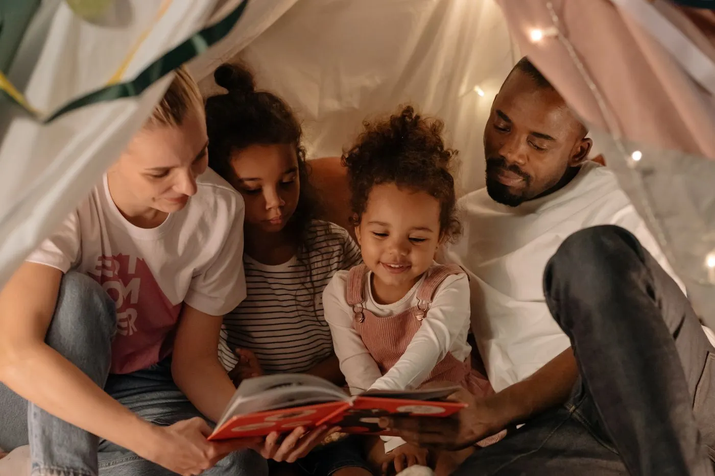 Família lendo um livro junta dentro de uma cabana de lençóis com luzinhas